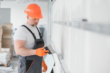 man drywall worker or plasterer putting mesh tape for plasterboard on a wall using a spatula and plaster. Wearing white hardhat, work gloves and safety glasses. Image with copy space. - obrazy, fototapety, plakaty