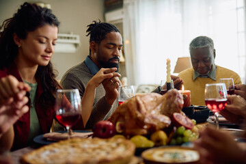 Black man and his family saying grace during Thanksgiving dinner at dining table.