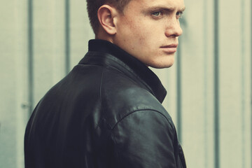 Tough guy concept. Handsome blue-eyed young man with short blond hair wearing black leather jacket, posing over urban corrugated background. Close up. Outdoor shot