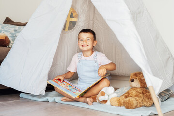 Portrait of smiling cute little preschooler boy reading book to his toys sitting in cozy kids...