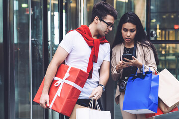 Multiracial couple standing outside mall after shopping and using smartphone