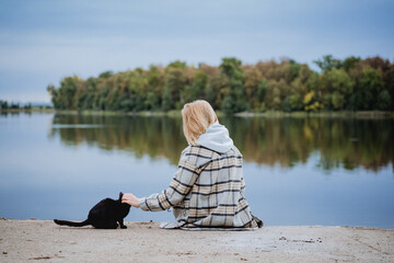 A girl in a plaid shirt sits on the pier and strokes a cat, a black cat caresses a person, love for...