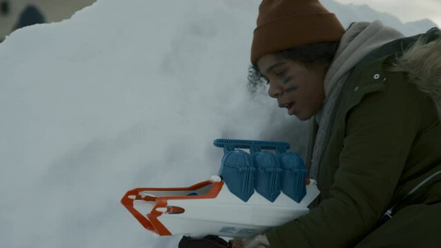 Medium handheld shot of young black girl in parka jacket and warm hat hiding behind snow wall, loading toy gun, standing up and firing snowballs at enemy fortress, while playing outdoors on winter day
