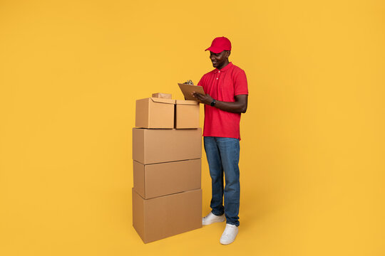 Busy focused millennial black guy courier in uniform making notes in warehouse, cheks many boxes