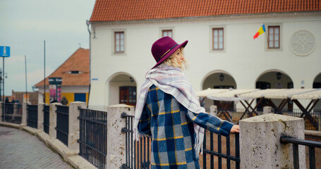 Fashioned woman wearing coat scarf and hat standing next to railing. Visitor getting acquainted...