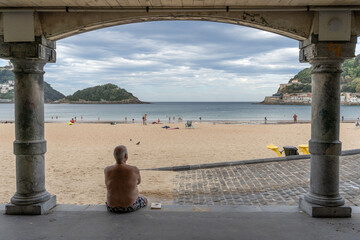 vue sur une plage depuis les arches d'un vieux bâtiment avec un vieux monsieur en maillot de bain assis de dos - 666753839