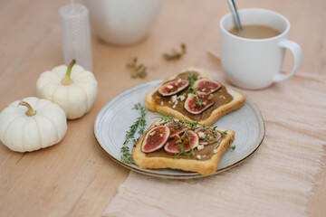 Home autumn breakfast, two toasts with almond butter, figs, coffee, pumpkins, candle on a round wooden table.
