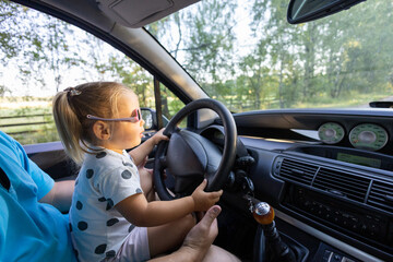 A cute little girl driving a car on her father's lap. Child girl in sunglasses with a serious...