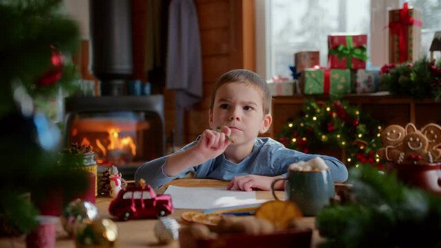 little diligent boy writes a letter with wish list to santa claus using color pencils. thinks over and says oh. christmas decorations, fir tree branches, flashing lights and fireplace on background