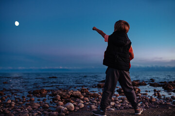 Boy standing on shore of lake at night and pointing to the moon - 666763020