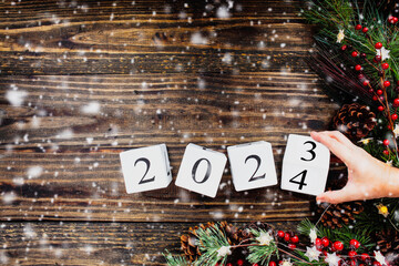 Woman's hand flipping New Year's 2023 wood calendar blocks to 2024. Christmas tree lights, pine...
