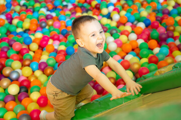Fototapeta na wymiar Happy smiling toddler boy climbs up on a slide in a children's room or children's entertainment center. Multi-colored balls. Happy active pastime and childhood. Playground. Selective focus