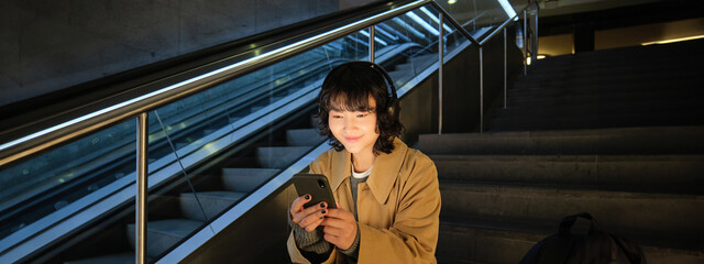 Technology. Portrait of asian girl sits on stairs near escalator, listens music in headphones and...