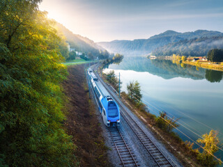 Aerial view of blue modern high speed train moving near river in alpine mountains in fog at sunrise...