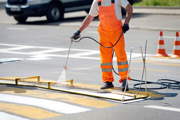 Worker painting zebra crossing stripes with spray gun. Road surface marking, painting and remarking pedestrian crosswalk. Worker use paint sprayer, applies white marking to striped crosswalk - obrazy, fototapety, plakaty
