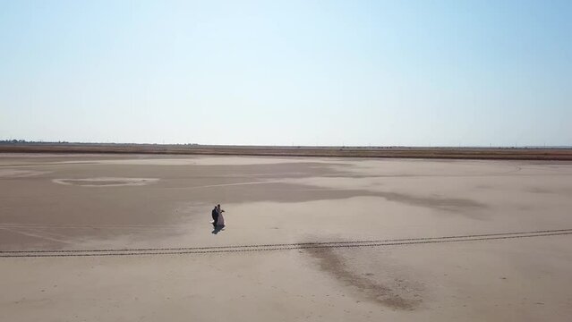 Aerial of newlyweds going at sandy desert landscape of dry lake. Bride in wedding dress, groom in suit walking at photoshoot. Just married man, woman enjoy wedding day. Drone video, global warming.