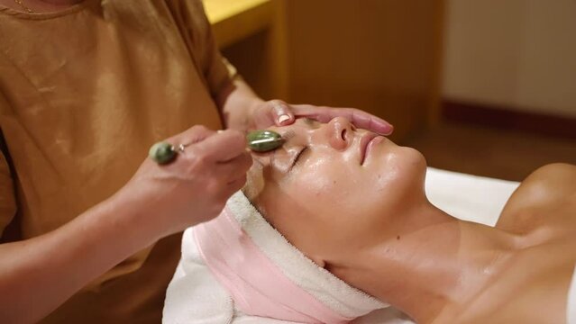 Young caucasian woman gets lifting facial massage with a rolling tool at spa and wellness center. Female client experiences facial lymphatic drainage therapy using jade roller and massage oil closeup
