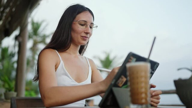 Female freelancer draws with stylus on a graphics tablet in cafe with ocean view, palm trees. Artist girl working on device by the seaside. Young illustrator on remote work day on tropical island.
