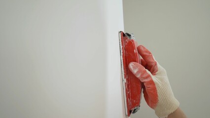 A man's hand sands a wall after plastering. A plasterer or painter sands white walls. Sanding and leveling the wall after applying putty. Sanding the wall after puttying using sandpaper.