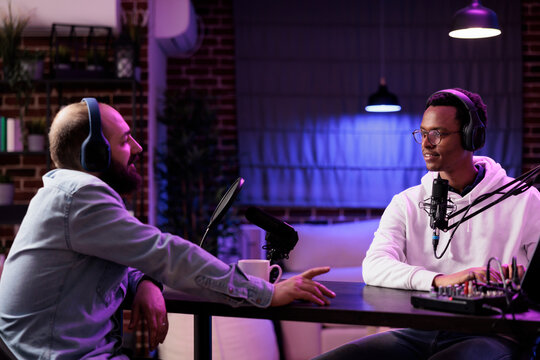 Influencer recording podcast with guest, using audio recording equipment in home studio living room. Cheerful men chatting live during broadcasting session for online streaming service show