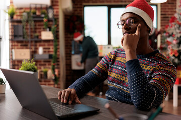 Startup company african american man employee in santa hat working on laptop in office at christmas holiday. Worker thinking while managing project report at festive decorated workplace - obrazy, fototapety, plakaty