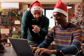 Two smiling african american and caucasian colleagues in santa hats collaborating on project in...