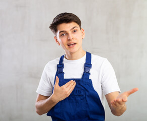 Young experienced worker in jumpsuit is actively gesticulating,pointing hands to side and inviting...