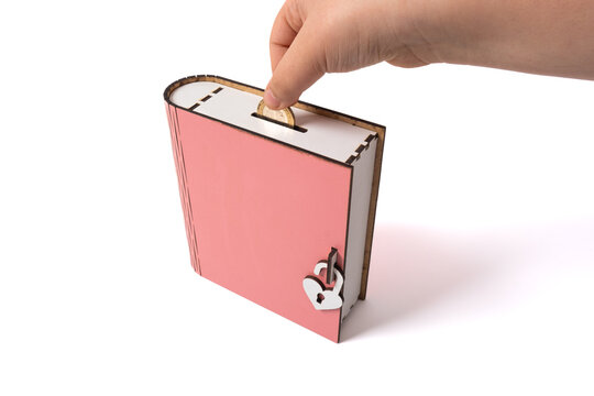 Girl puts money into pink piggy bank money box in the form of a book on white background. Financial literacy concept for children. Coin savings, top view, isolated