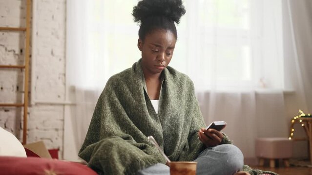 Sad young woman under the blanket hold ice cream while looking at smartphone reading bad news break up message from ex-boyfriend or personal problems at home Upset brunette crying indoors