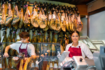 Positive young female and male vendors working at counter in butcher shop, arranging dry-cured...