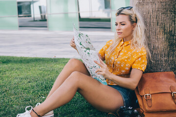 Blond lady sitting under tree with sunglasses on head and studying map