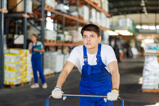 Young loader guy in overalls rolls cargo cart in warehouse