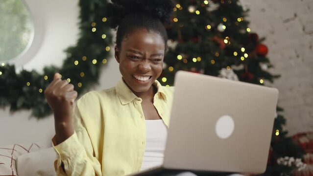 Amazed young woman extremely happy after read message on laptop computer at home Excited girl pleased rejoices received great successful news or result near Christmas tree at holidays indoors
