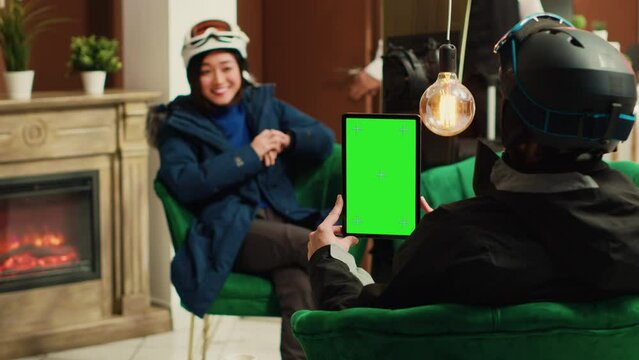 Traveller shows isolated greenscreen on tablet while she sits with skiing partner in lounge area, preparing for winter sport with gear. Man holding gadget with blank chromakey mockup template.