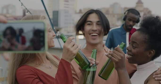 Group of friends with beer bottles posing for smartphone camera enjoying rooftop party celebrating event together. Professional DJ working in background.