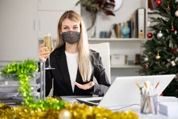 Business woman in face mask sitting at the desk with glass of champagne, celebrating New Year