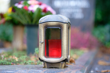 close up of brass grave lamp with red candle and with bouquet of flowers and gravestone in blurred...