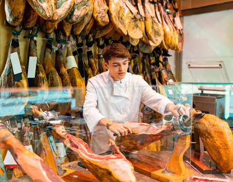 Smiling young guy seller in white uniform working at counter in butcher store, cutting slices from whole leg of dry-cured Iberian jamon on jamonera