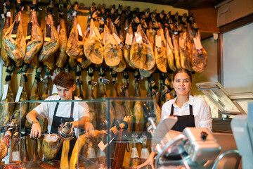 Young woman and man in uniform cutting ham with special knife. Woman and man selling traditional Spanish dish jamon.