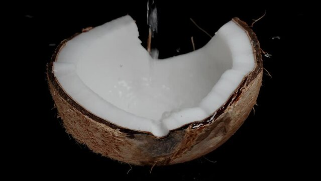 Splashes of coconut milk, juice dripping into a half coconut rotating on a black background, in slow motion.