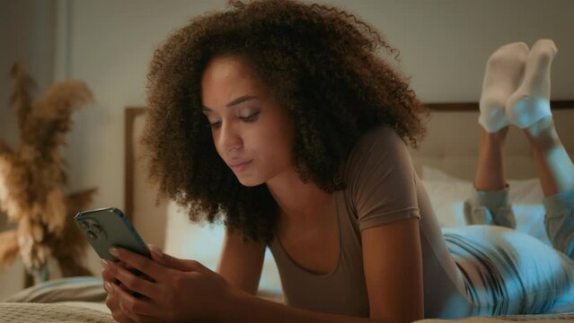 Upset African American woman ethnic girl teen gen z user scrolling mobile networks lying on bed in evening night bedroom phone chatting read bad news on smartphone device scam message failure error