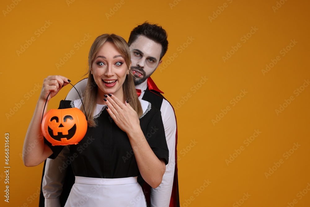 Poster couple in scary maid and vampire costumes with pumpkin bucket on orange background, space for text. 