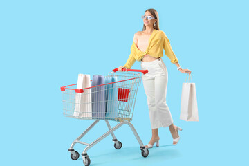 Stylish happy young woman with shopping bags and cart on blue background