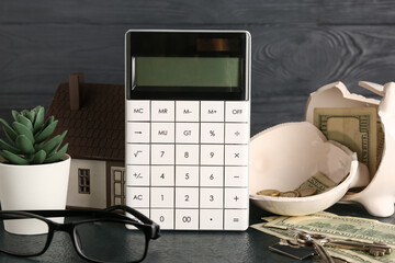 Calculator, broken piggy bank, money and house model on black table