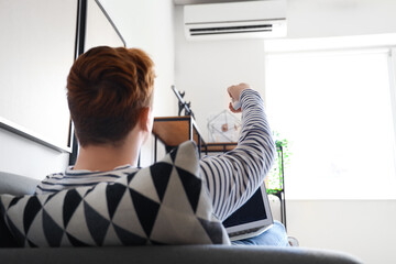 Young man with laptop switching on air conditioner at home, back view