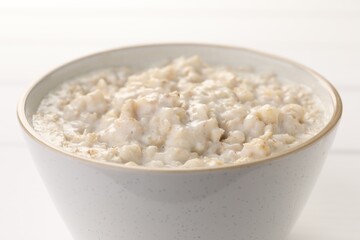 Tasty boiled oatmeal in bowl on white table, closeup