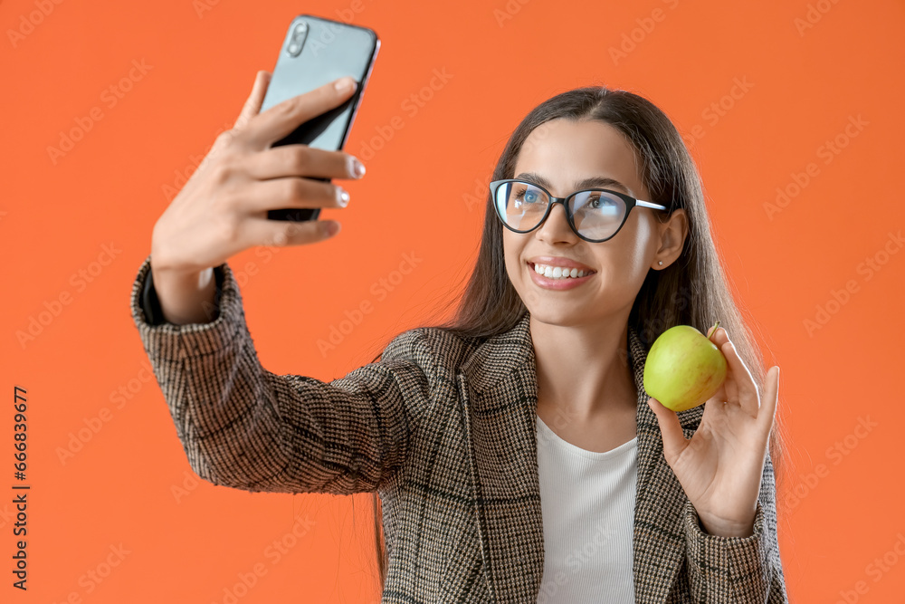 Poster Pretty young woman taking selfie with ripe apple on orange background