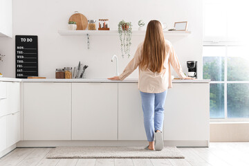 Back view of young woman standing near white counters in modern kitchen - obrazy, fototapety, plakaty