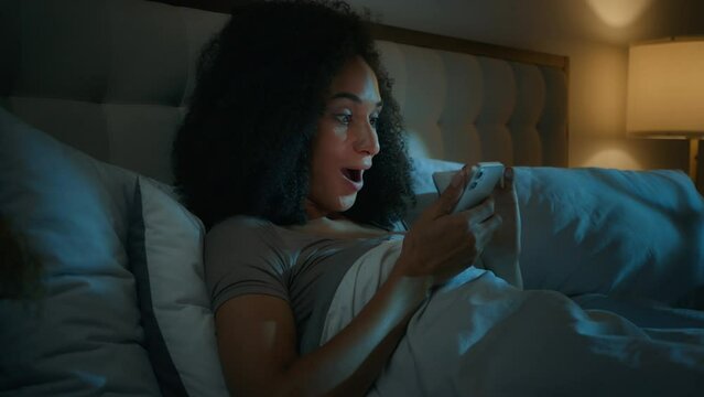 African American young woman girl lying in bedroom dark night insomnia gadget addicted playing online game with mobile phone on bed happy excited female winner winning achieve success victory prize