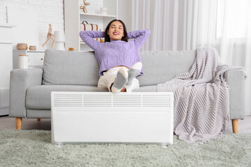 Pretty young woman relaxing on sofa with electric heater in living room